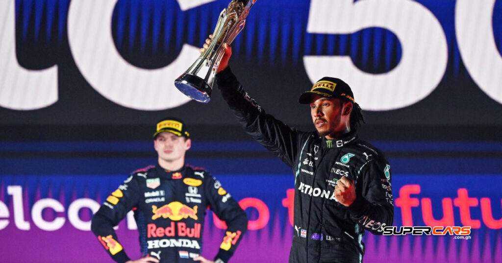 Lewis Hamilton celebrating his victory on the podium after winning a race in the 2024 Formula 1 season, holding his trophy with a wide smile, while his Mercedes team cheers in the background.