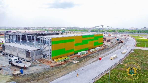 Construction site at Borteyman Sports Complex, showing the development of a new facility with vibrant green and yellow exterior panels, along with surrounding infrastructure work.