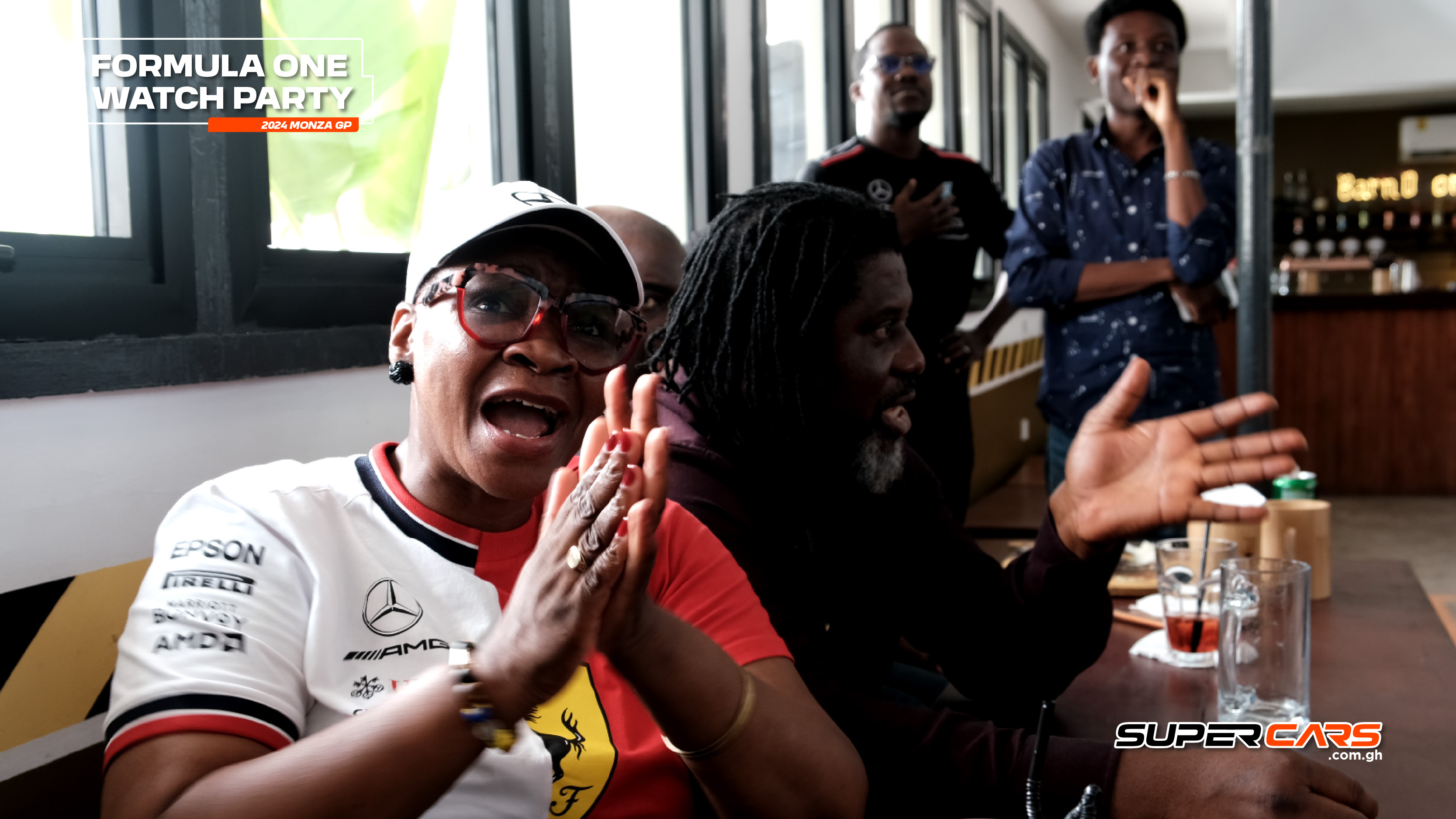 A lively Formula One watch party scene, showcasing an excited group of fans. A woman in a Ferrari shirt passionately cheers and claps, while others engage in the excitement around her. The atmosphere is vibrant, filled with enthusiasm for the 2024 Monza GP.