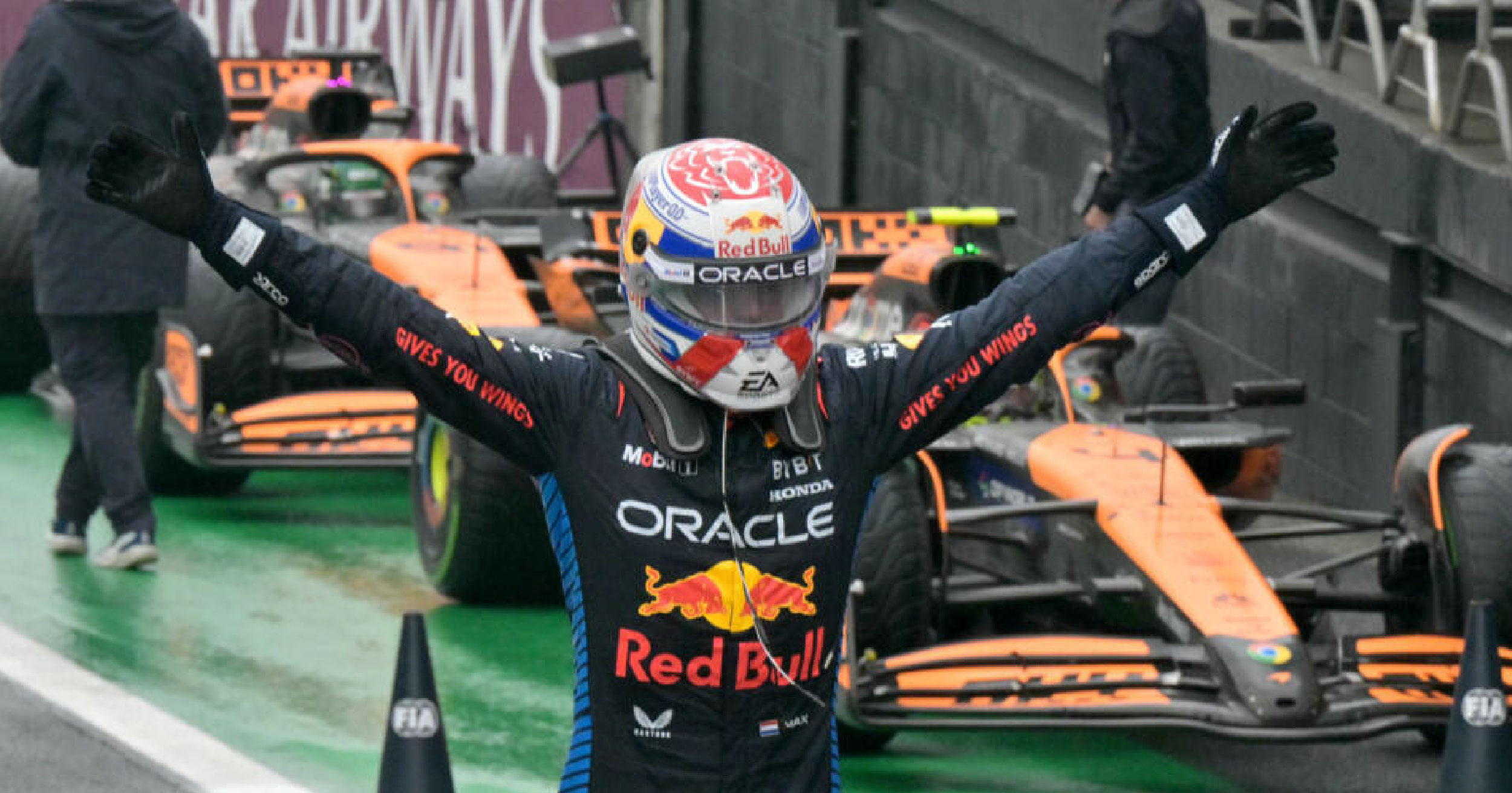 Max Verstappen raising his arms in celebration after winning the Brazilian Grand Prix, with Red Bull racing cars in the background.