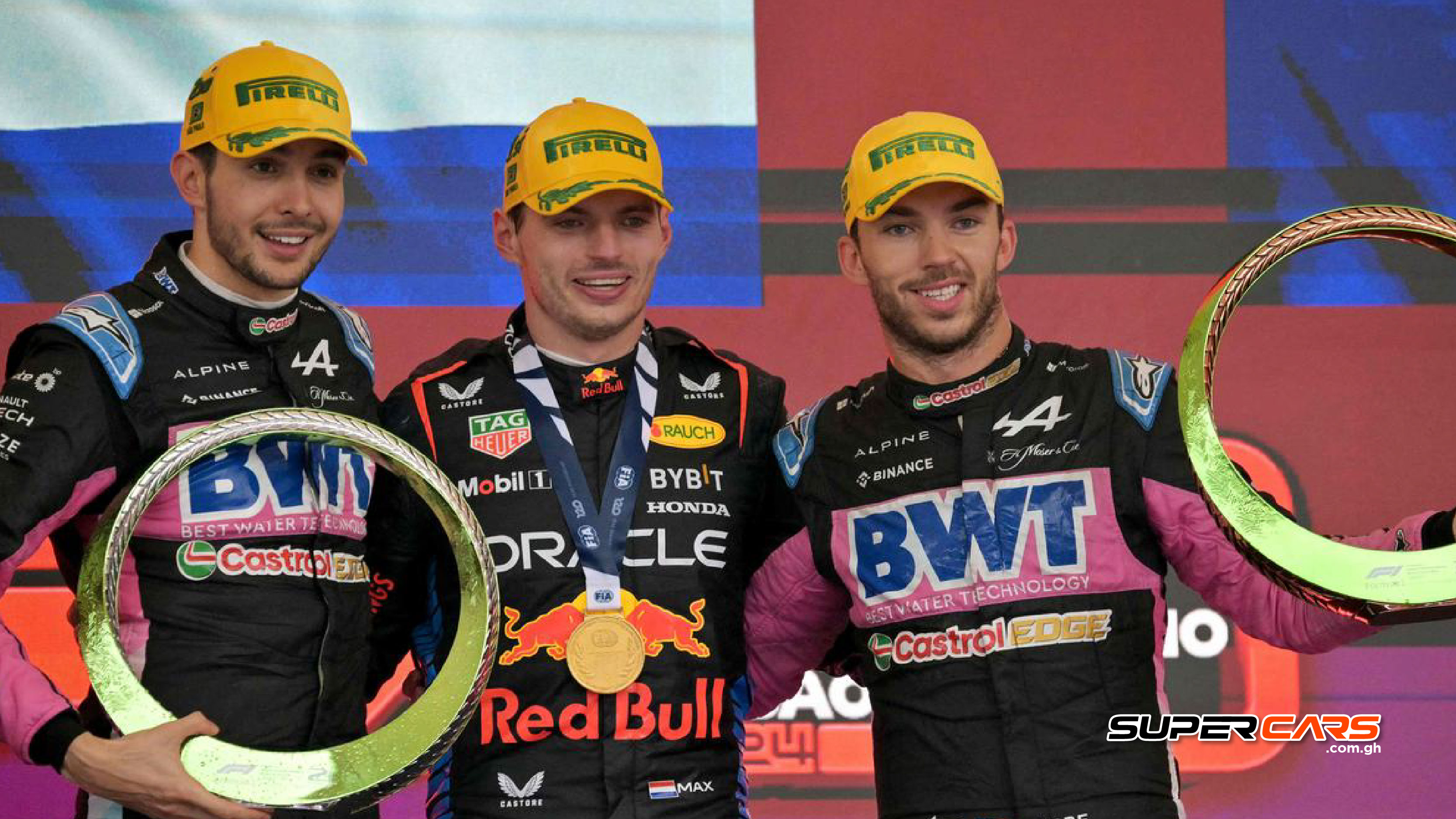 Esteban Ocon, Max Verstappen, and Pierre Gasly smiling on the podium with their trophies after the Brazilian Grand Prix, all wearing Pirelli hats and their team race suits.