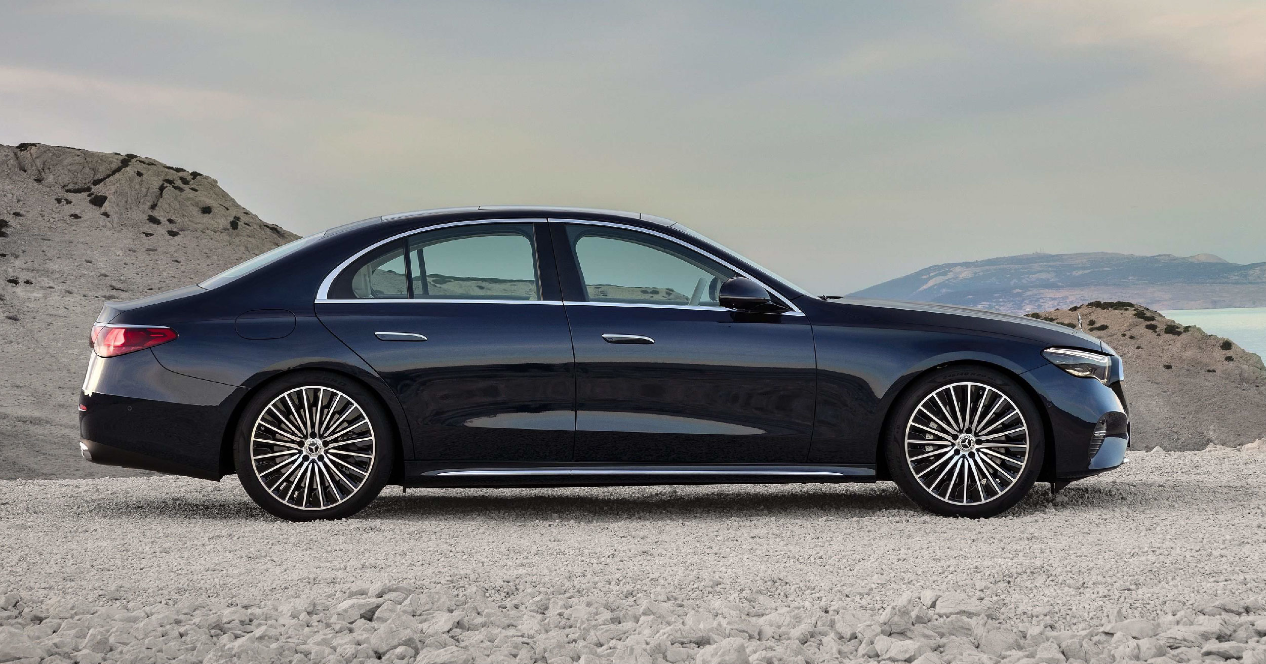 Side profile of a dark blue Mercedes-Benz E-Class sedan parked on a rugged landscape with mountains in the background, emphasizing its sleek and refined silhouette.